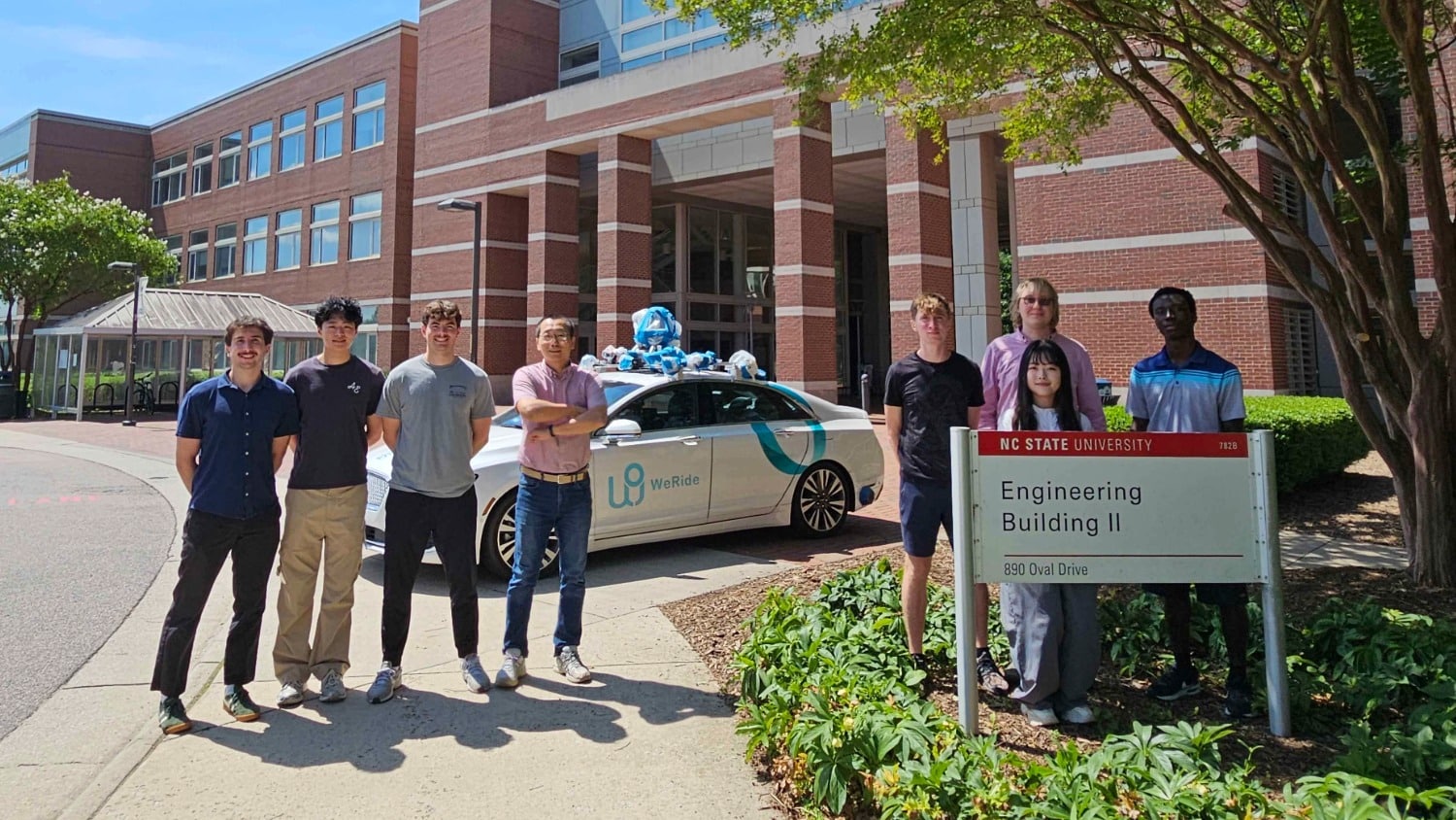 A small group of people stand in the sunshine outside a large academic building in front of an autonomous driving vehicle.